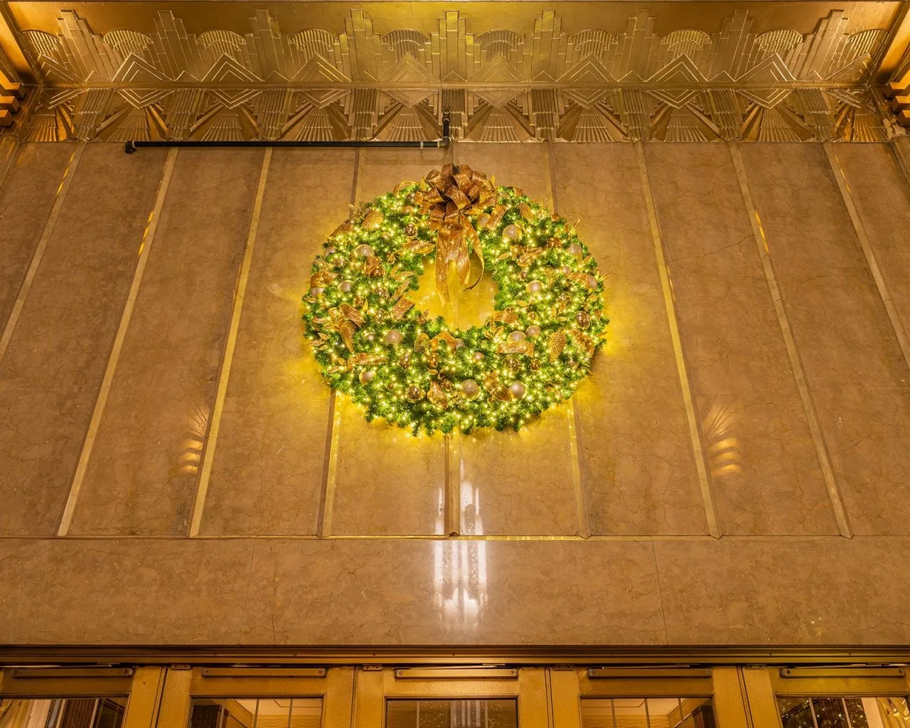 An illuminated wreath hangs high on a paneled stone wall above a lobby doorway.