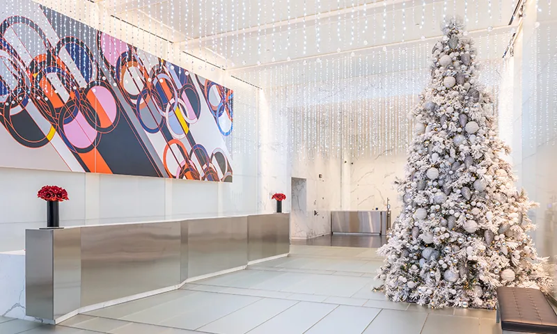 A snow-toned tree stands beside a long reception desk beneath hanging lights in a bright lobby.