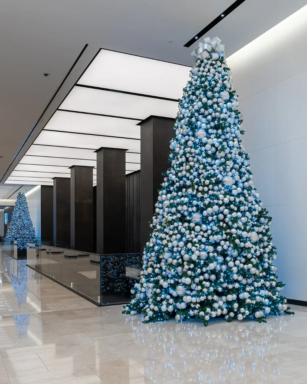 Silver and white ornaments on illuminated trees beside dark columns in a bright lobby