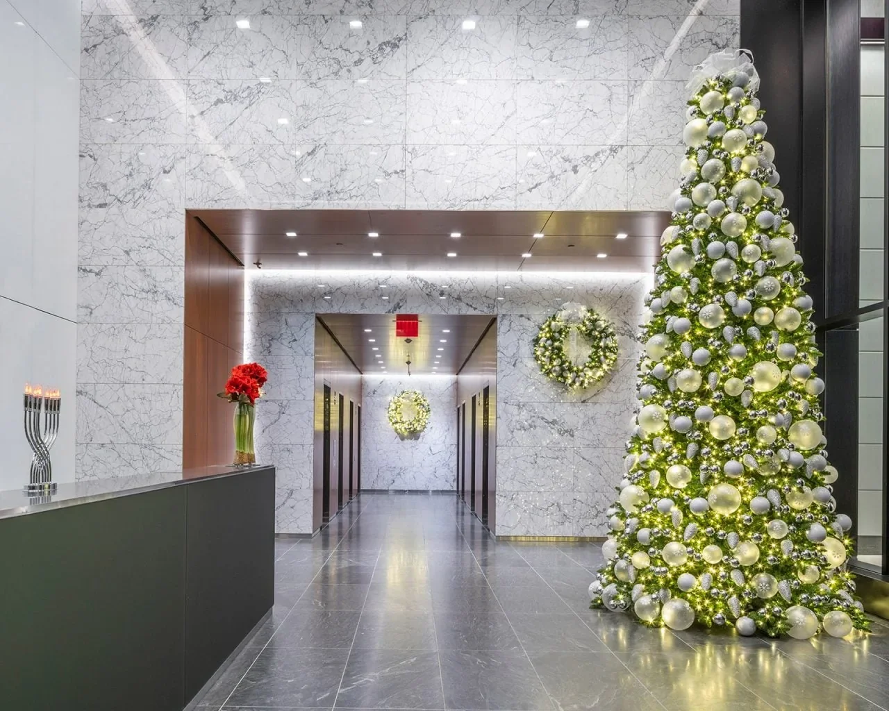 Tall decorated tree, illuminated wreaths and a menorah in a bright marble lobby with a reception desk